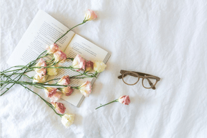 A photo of an open book on a bed with white and pink roses piled on top. Reading glasses sit next to the book.