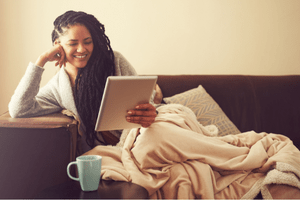 A woman smiles at the tablet she is holding and she lounges on a couch with a blanket and a warm beverage