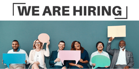 A diverse group of individuals sit on chairs next to one another in a line, holding up speech bubble props, laughing and smiling for a photo. Text says, "we are hiring".