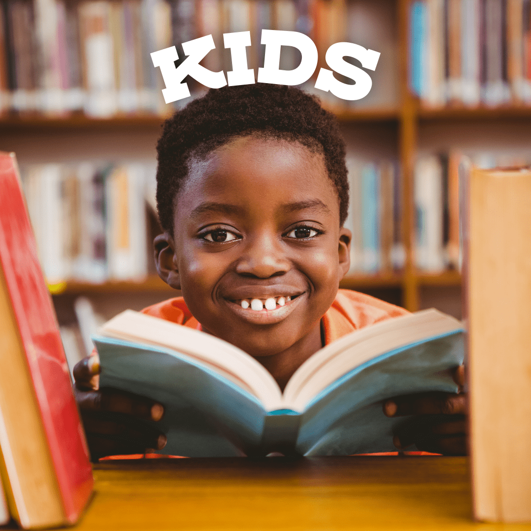 A Black boy holding a book smiles behind a shelf of books